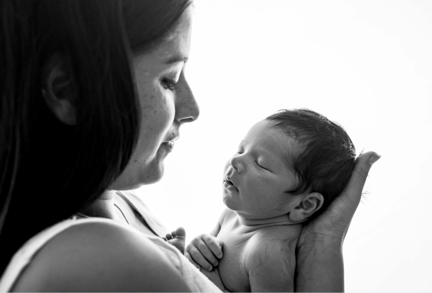 Black-and-white close-up of a mother gently holding her sleeping newborn, gazing at the baby as she supports the baby’s head with one hand.